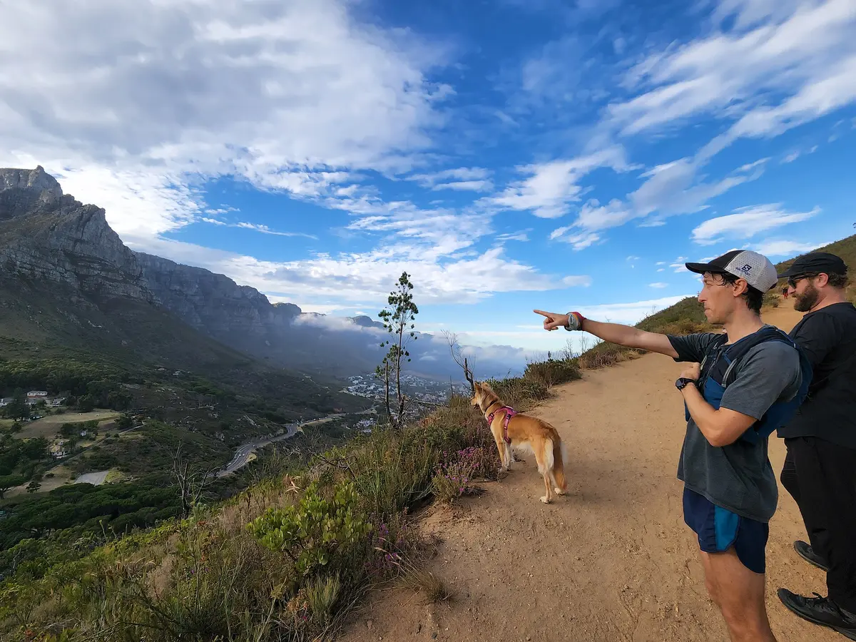Two hikers and a tan dog in a harness on a mountain path high above a green valley, under a bright blue sky