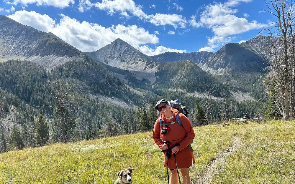 A hiker and her dog on a green mountain meadow trail under a blue summer sky